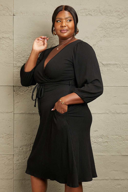 Plus size woman wearing a black Culture Code full size surplice flare ruching midi dress with long sleeves and waist tie