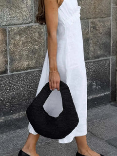 Woman wearing a white ruffled V-neck maxi dress holding a black textured handbag walking on a stone pavement