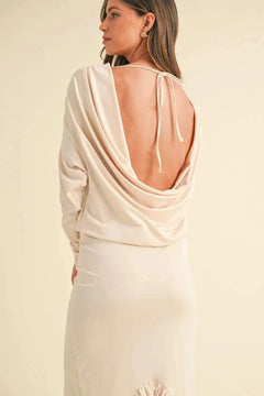 Woman wearing a cream backless asymmetric ruffle hem dress with deep cowl neck and tied back detail.
