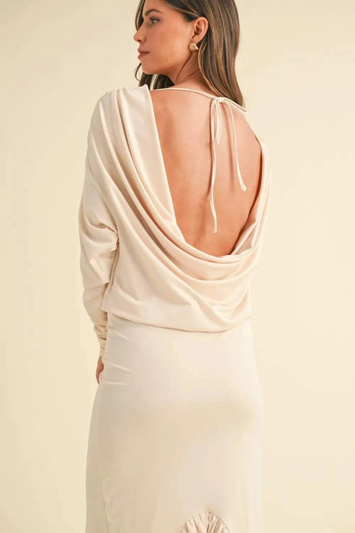 Woman wearing a cream backless asymmetric ruffle hem dress with deep cowl neck and tied back detail.