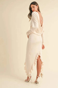 Woman wearing a beige backless asymmetric ruffle hem dress with long sleeves and high heels