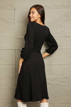 Woman wearing a black Culture Code Full Size surplice flare ruching midi dress with long sleeves and waist tie viewed from the back
