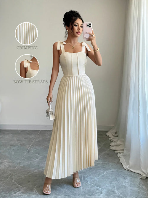 Woman wearing cream pleated tie-shoulder maxi dress with bow straps and square neckline, posing indoors with small handbag and transparent heels.