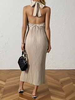 Back view of a pleated halter neck midi dress in beige with tied bow detail and no lining, accessorized with a black handbag and white heels.
