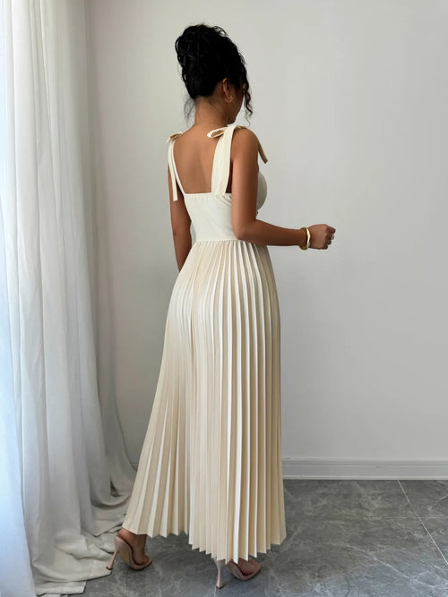 Woman wearing a cream pleated tie-shoulder maxi dress with bow details and no stretch, standing indoors
