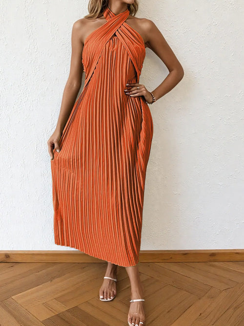Woman wearing an orange pleated halter neck midi dress with crisscross ties and no stretch, styled with sandals on wooden floor.