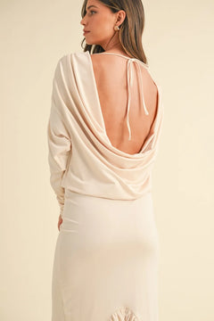 Woman wearing a cream backless asymmetric ruffle hem dress with deep cowl neck and tied back detail.