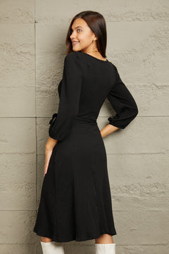 Woman wearing a black Culture Code Full Size surplice flare ruching midi dress with long sleeves and waist tie viewed from the back